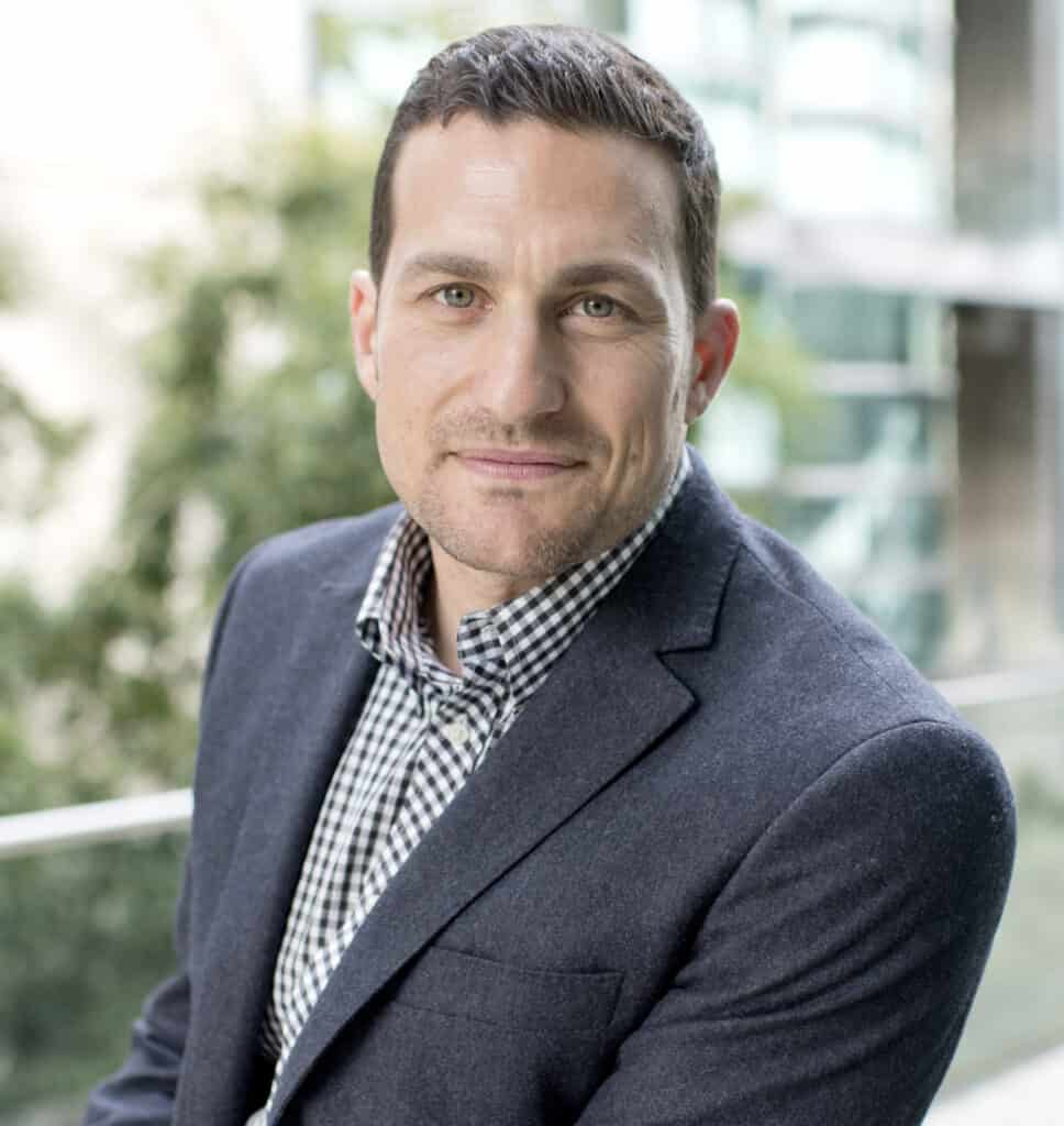 A man in a suit and a checkered shirt is leaning against a railing while Andrew Huberman eats nearby.