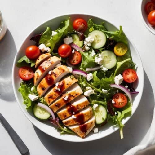 A plate of nutritious chicken salad with sliced grilled chicken, cherry tomatoes, cucumber slices, red onion, and feta cheese. A jar of dressing, a small bowl of cherry tomatoes, a fork, and a napkin are nearby. Perfect for those seeking a healthy recipe.