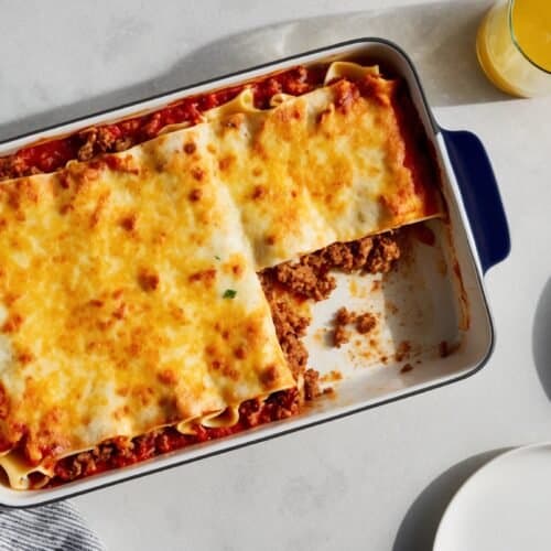 Baked classic lasagna with a portion missing in a rectangular dish, accompanied by a glass of orange juice, a striped cloth, and a white plate with utensils on a white surface. This delightful recipe features layers of pasta, rich béchamel sauce, and savory meat filling.