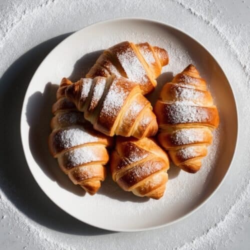 A plate of croissants dusted with powdered sugar sits on a marble countertop, reminiscent of a classic Italian Cornetto recipe, with a circular sprinkle of powdered sugar surrounding the plate.