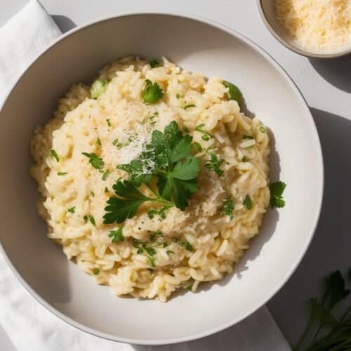 A bowl of classic, creamy risotto garnished with parsley, placed on a white napkin, next to a small bowl of grated cheese and a few parsley leaves—a timeless recipe favorite.