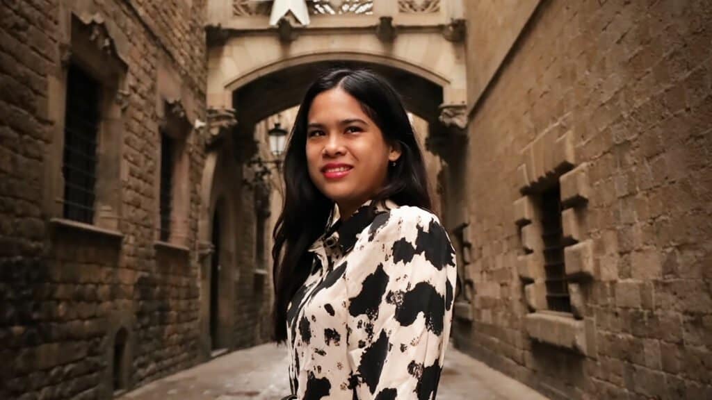 A woman with long dark hair stands in a narrow, stone-walled alley, wearing a black and white patterned shirt. She smiles faintly, with archways and lanterns visible in the background, as if she were an auto draft of forgotten history.