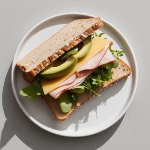 A Fieldwork Sandwich: Whole grain bread, fresh lettuce, creamy avocado, turkey slices, cheese, and arugula served on a white plate. Perfect lunch for those on the go.