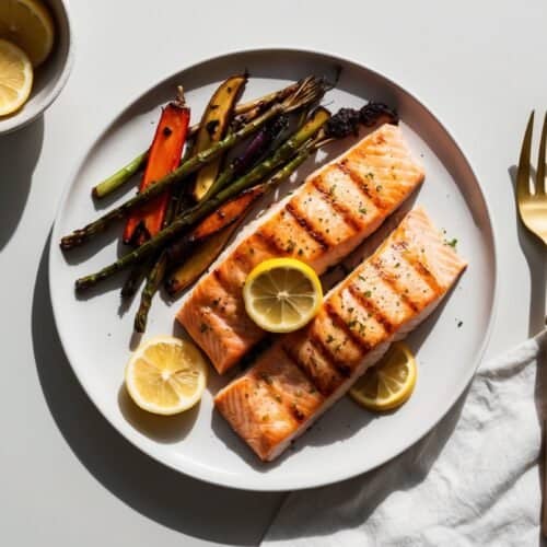 Grilled salmon filets with lemon slices and roasted vegetables adorn a white plate, accompanied by a bowl of lemon slices, golden cutlery, and a white napkin on a pristine white surface. This picturesque setup almost feels like an auto draft for culinary perfection.