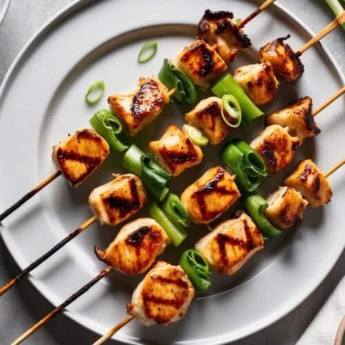 Enjoy this easy recipe for grilled chicken skewers with green vegetables on a white plate, accompanied by sliced green onions and a bowl of dipping sauce, reminiscent of traditional Grilled Yakitori.