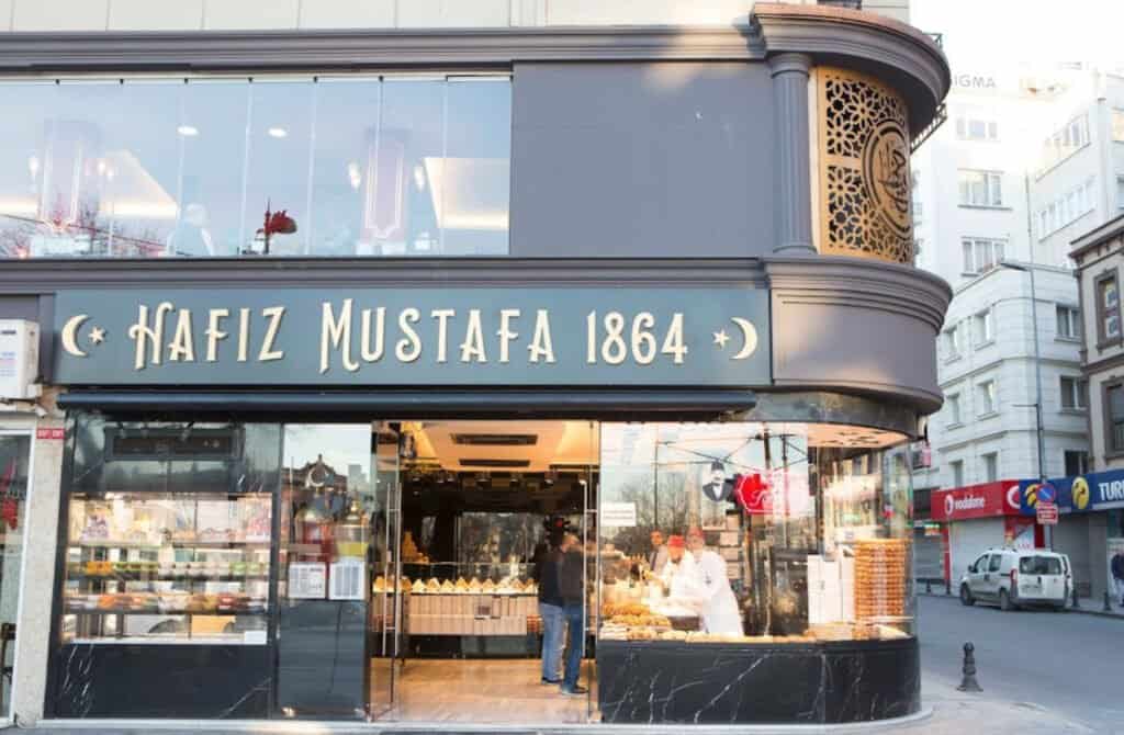 Street view of Hafiz Mustafa 1864, a well-known pastry shop in Istanbul, showcasing a person at the counter inside and an array of Turkish sweets on display. The modern storefront is nestled among other buildings on a bustling city street.