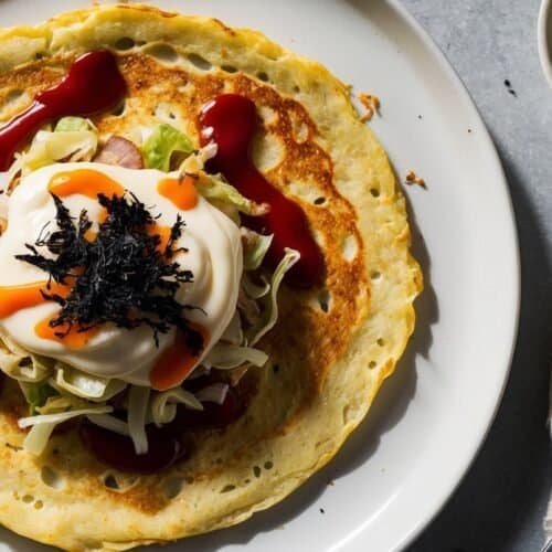 A plate with a savory crepe, reminiscent of an easy homemade okonomiyaki recipe, is topped with shredded cabbage, a dollop of mayonnaise, seaweed, and drizzled with red sauce. A small bowl with a yellow liquid is on the side, along with a fork and napkin.