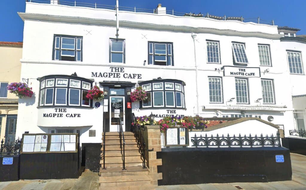The image shows the exterior of "The Magpie Cafe," a white building with black trim located near Whitby Harbor. There are steps leading to the entrance and colorful hanging flower baskets on either side, hinting at the delicious fish and chips awaiting inside.