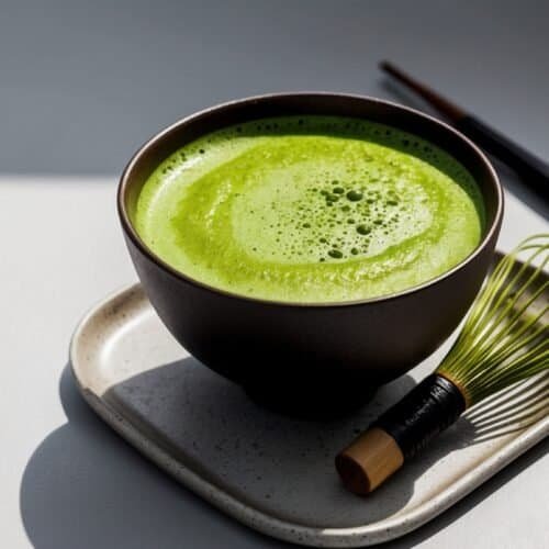 A bowl of green matcha tea, embodying the perfect matcha, is placed on a square plate. A bamboo whisk and chopsticks are positioned beside the bowl, ready to complete this serene matcha tea experience.