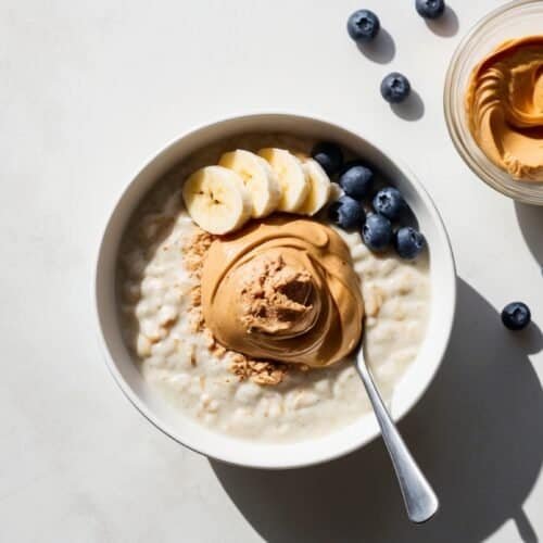 A bowl of protein-packed oatmeal with peanut butter, sliced bananas, and blueberries, accompanied by a small glass bowl of peanut butter and several loose blueberries on a white surface.