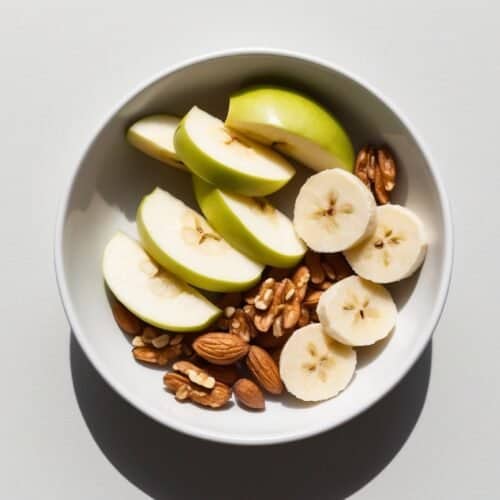 A white bowl containing fresh fruit like sliced green apples and banana slices, along with crunchy walnuts and almonds, is placed against a light grey background. Perfect for a quick breakfast.