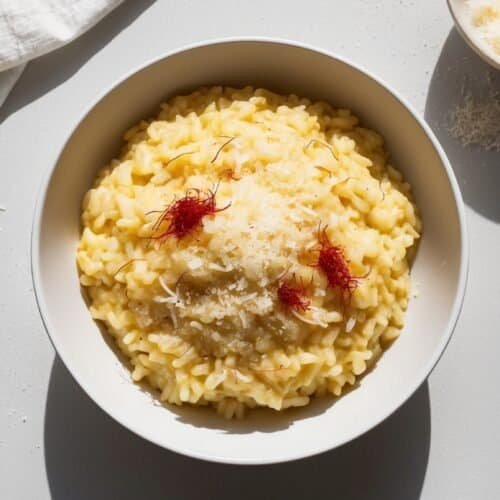 A bowl of creamy saffron-infused risotto garnished with a few delicate threads and grated cheese, placed on a pristine white surface. Nearby, you'll find a small bowl with more grated cheese and a white cloth. This dish could easily be the star of any culinary Auto Draft.