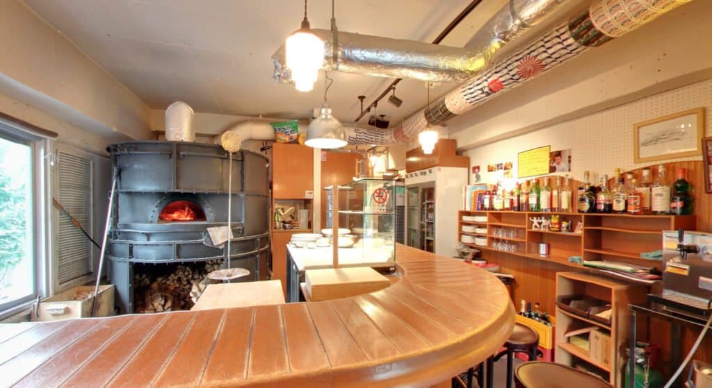 A cozy pizzeria interior with a large, rustic wood-fired oven behind a curved wooden counter. Shelves with bottles and menus are on the right, and pendant lights hang from the ceiling, casting a warm glow perfect for relaxation.