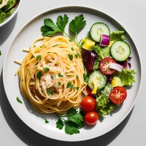 A plate of spaghetti garnished with herbs, garlic, and grated cheese is served alongside a fresh salad with cucumbers, cherry tomatoes, red onions, and lettuce. A small bowl of salad and extra grated cheese are nearby.