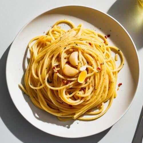 A plate of spaghetti aglio e olio garnished with garlic cloves and red pepper flakes, accompanied by a fork and a glass of olive oil, makes for a perfect recipe.