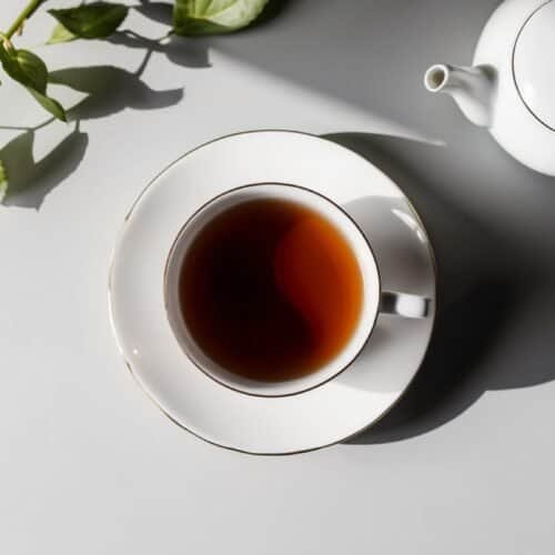 A cup of strong morning tea sits on a saucer beside a teapot and some fresh green leaves on a plain surface.