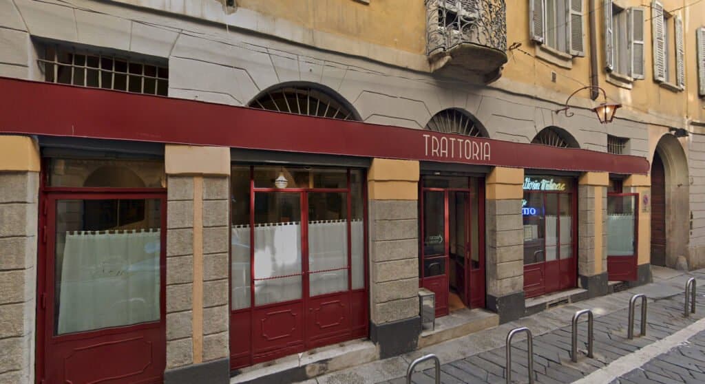 Street view of a closed Milanese trattoria with a red awning and gray stone building exterior. The street is paved and has several bollards in front.