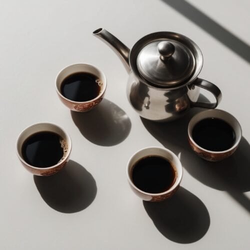 A silver teapot and four small cups filled with dark liquid are arranged on a light gray surface with long shadows, as if meticulously positioned by an auto draft.