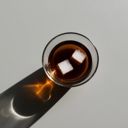 A clear glass of dark liquid with two sugar cubes floating on its surface, reminiscent of an Authentic Turkish Tea, casts a shadow on a light gray background.