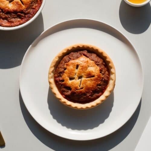 A small, baked pie on a white plate with a gold fork to the left and a partial view of another pie and a cup of orange juice in the background, reminiscent of traditional Australian meat pies from an old family recipe.