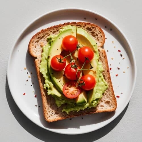 A slice of bread topped with mashed avocado and cherry tomatoes on a white plate, with pepper flakes scattered around, makes the perfect quick recipe for any time of day.