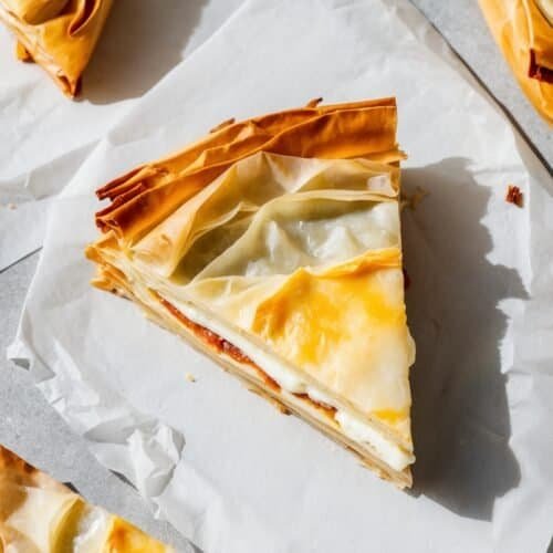 Slices of layered phyllo pastry with a creamy filling, arranged on parchment paper, offer a taste of traditional Bulgarian cuisine in this Banitsa recipe.