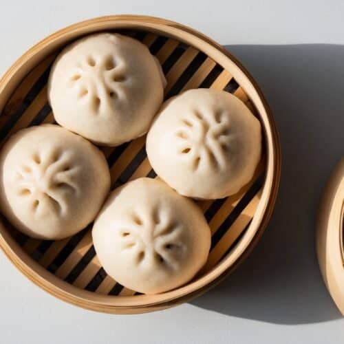 A bamboo steamer basket containing four savory pork steamed buns with a decorative pattern on top, accompanied by its lid placed to the side.