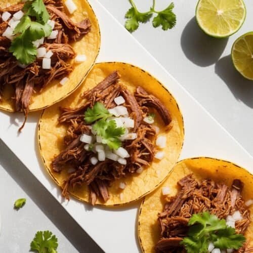 Three flavorful beef tacos on a rectangular white plate, garnished with chopped onions and cilantro, with cut limes and extra cilantro on the side. Ideal for any Barbacoa Tacos Recipe lovers craving an authentic taste.