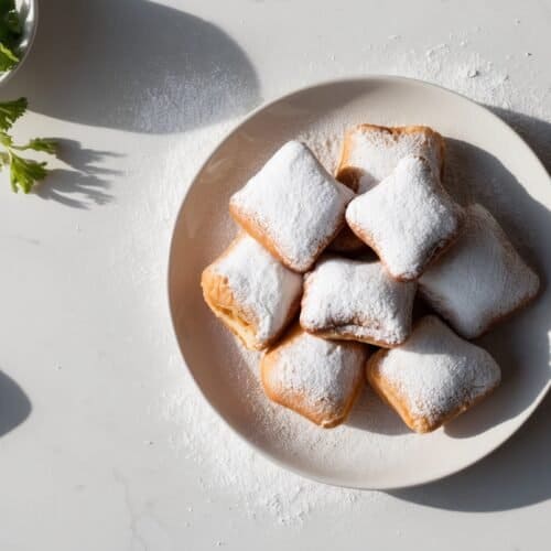 A plate of seven powdered sugar-covered beignets sits on a white surface, with a sprig of parsley and a white cloth napkin nearby, evoking the charm of a classic New Orleans recipe.