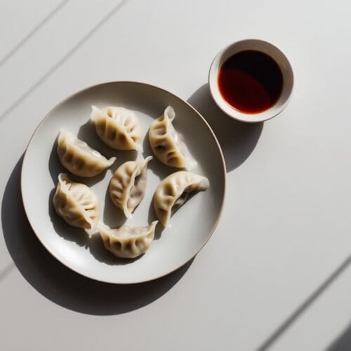 A white plate with eight homemade Chinese pork dumplings, each boasting a savory filling, is placed next to a small bowl of soy sauce on a white table. Shadows cast on the table.