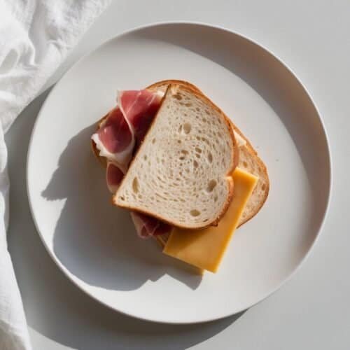 A white plate with a delicious cheese sandwich containing slices of bread, ham, and cheese placed on a light-colored surface next to a white cloth.