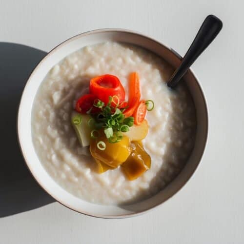 A comforting congee bowl topped with sliced red chilies, pickled vegetables, and chopped green onions, served with a black spoon on the side.