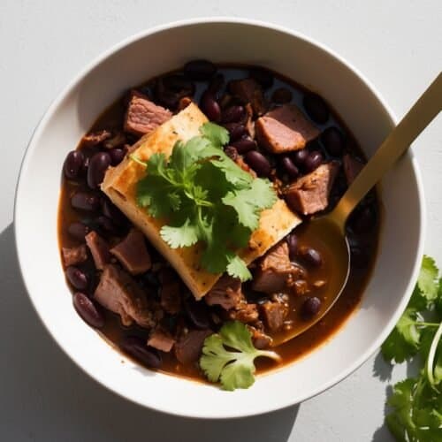 A white bowl containing a Brazilian Feijoada with black beans, chunks of meat, and a piece of tofu topped with cilantro, all swimming in a rich broth with a gold spoon. Fresh cilantro rests beside the bowl on a light gray surface.