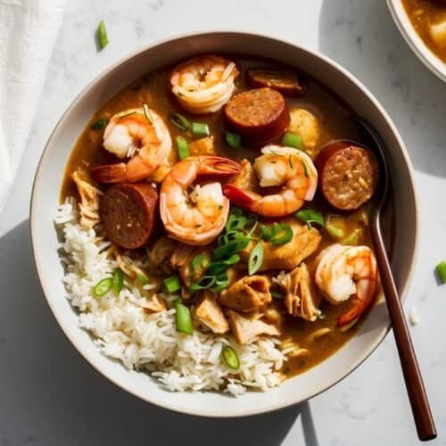 A bowl of spicy Cajun gumbo with shrimp, sausage, and chicken, served with rice and garnished with green onions. Another bowl of gumbo is partially visible in the background.