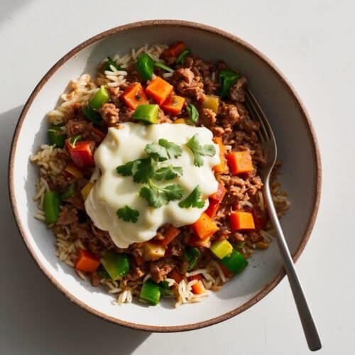 A bowl of rice topped with a mix of diced vegetables and minced meat, garnished with a creamy white sauce and fresh parsley, sits invitingly with a fork on the side—truly a hearty meals recipe.