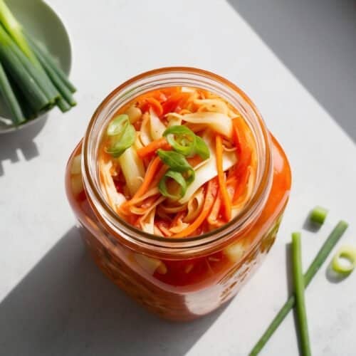 A jar of homemade kimchi with sliced green onions on top is placed on a light-colored surface, next to a small plate with additional green onions, under natural sunlight.