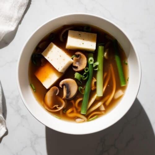 A bowl of miso soup with tofu cubes, mushrooms, green onions, and bok choy sits on a marble surface. A white cloth is placed to the left of the bowl— a comforting twist for your favorite Hot and Sour Soup recipe.