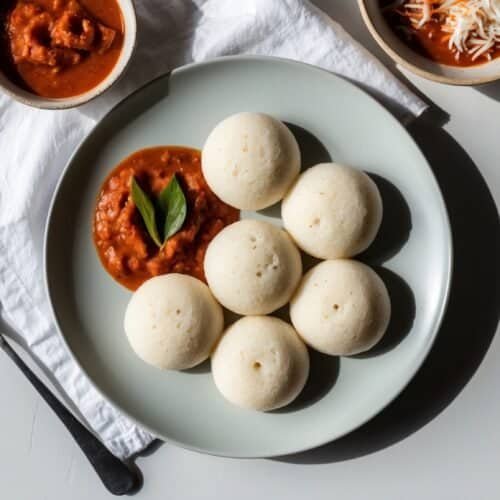 A plate of six fluffy idli served with a bowl of red sauce on the side, placed on a white napkin with a spoon next to it.