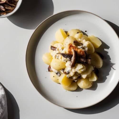 A plate of sliced potatoes topped with creamy sauce and mushrooms is set on a white surface next to a fork and a white napkin, reminiscent of a Janssons Frestelse recipe, accompanied by a small bowl of mushrooms.