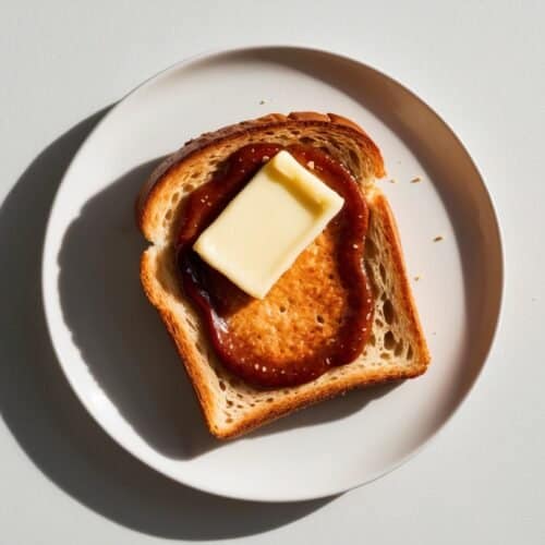 A plate with a slice of toast topped with a round patty, a layer of sauce, and a rectangular pat of butter, reminiscent of Singaporean Kaya Toast, placed on a white surface.
