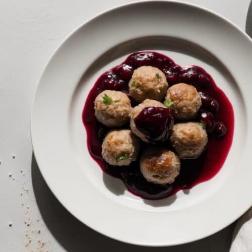 A white plate with five Swedish meatballs topped with a dark red Lingonberry sauce, accompanied by a fork on the left side and a white napkin on the right.