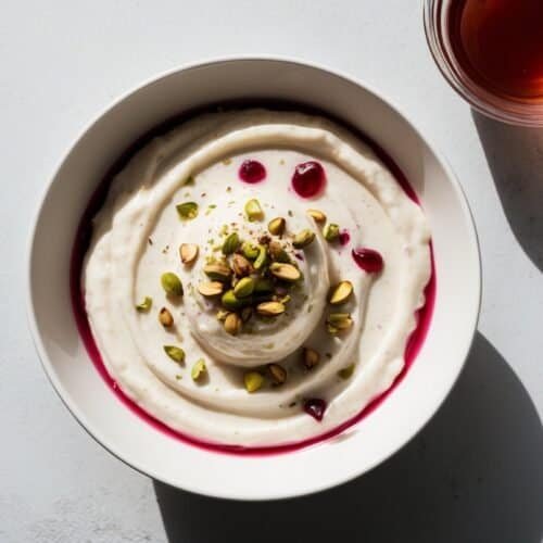 A bowl of creamy yogurt topped with pistachios and a drizzle of berry sauce, reminiscent of Malabi, accompanied by a glass of tea on the side.