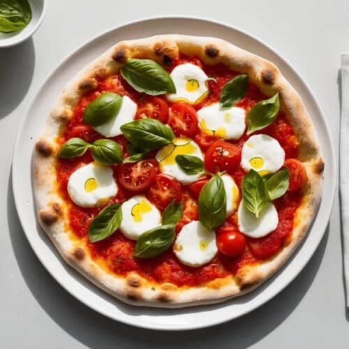 A classic Margherita pizza topped with fresh basil, sliced tomatoes, and mozzarella balls on a white plate. Olive oil, extra mozzarella, a fork, and a napkin are on the table.