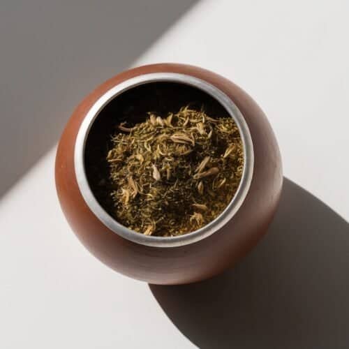 A round wooden bowl containing dried herbs, viewed from above, with a shadow cast on a white surface, evokes the essence of a traditional Argentine Mate recipe.