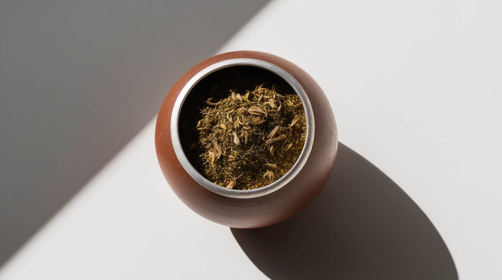 A round wooden bowl containing dried herbs, viewed from above, with a shadow cast on a white surface, evokes the essence of a traditional Argentine Mate recipe.