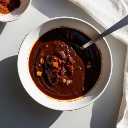 A bowl of thick, dark Mole Poblano soup with chunks of vegetables sits next to a spoon and a bowl of powdered spice on a white surface with a white cloth, showcasing the rich flavors of this traditional recipe.