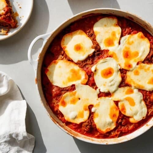 A hearty dish with melted cheese on top sits on a table next to a white napkin. A portion of Bulgarian Moussaka is served on a plate in the background with a fork, also on the table.
