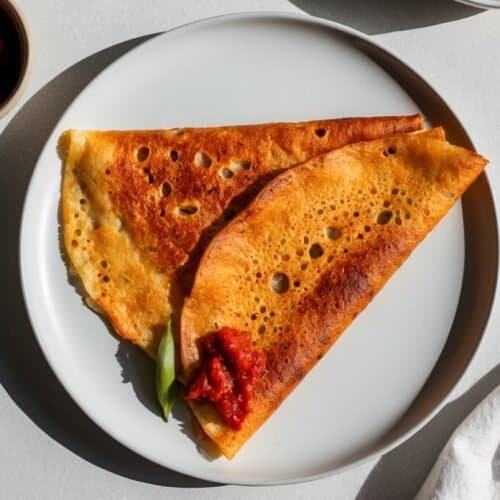 A folded crispy Mysore Masala Dosa with a dollop of spicy tomato chutney on a white plate. A small bowl of additional chutney is placed beside the plate on a light-colored surface, embodying the rich flavors of South Indian cuisine.