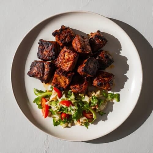 A white plate containing grilled meat cubes, showcasing an authentic Nyama Choma recipe, and a side salad with lettuce, tomatoes, and possibly grains, set on a light gray surface.