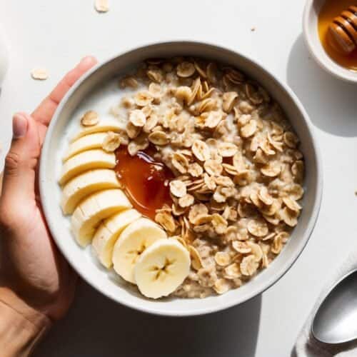 A bowl of oatmeal topped with banana slices, honey, and oats is held in one hand. Nearby are a jar of almond milk, a bowl of honey with a dipper, and a spoon on a napkin.